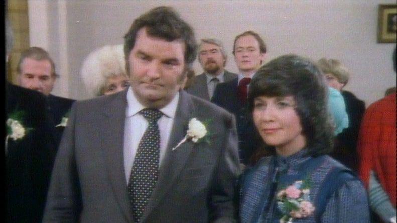 Lisabeth Miles as Megan Harries, wearing a blue dress with a floral cortege, on her wedding day in 1988, she is stood next to a man in a dark grey suit. There are people behind them. It is their wedding day with guests stood with them.