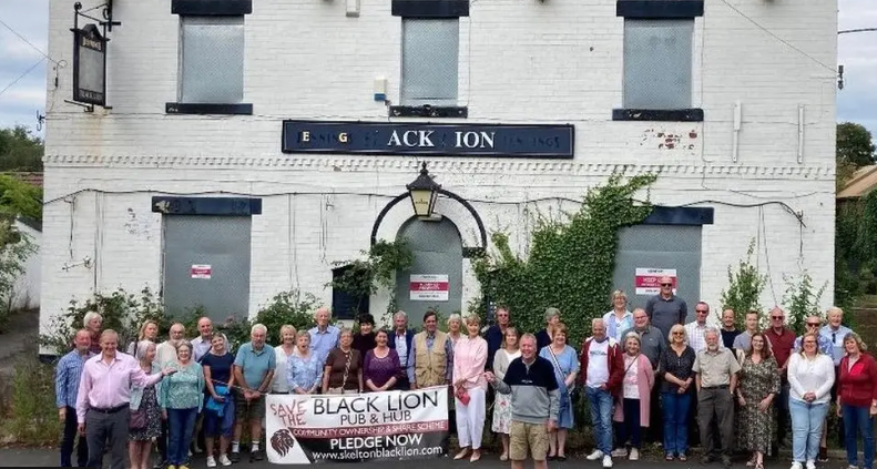 A mixed group of village residents stand outside the Black Lion pub before renovation. The building has been painted an off-white, and has ivy growing around the door, with metal shutters at the window.