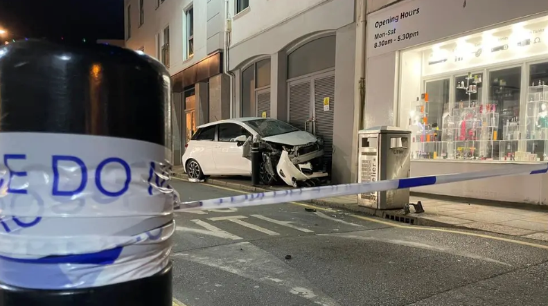 A white car with a damaged front end next to a shop with police tape crossing a road in the foreground.