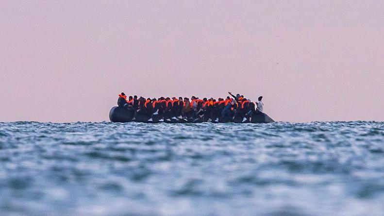 A side view of a small rubber boat filled with people wearing life jackets crosses the English Channel towards the UK.
