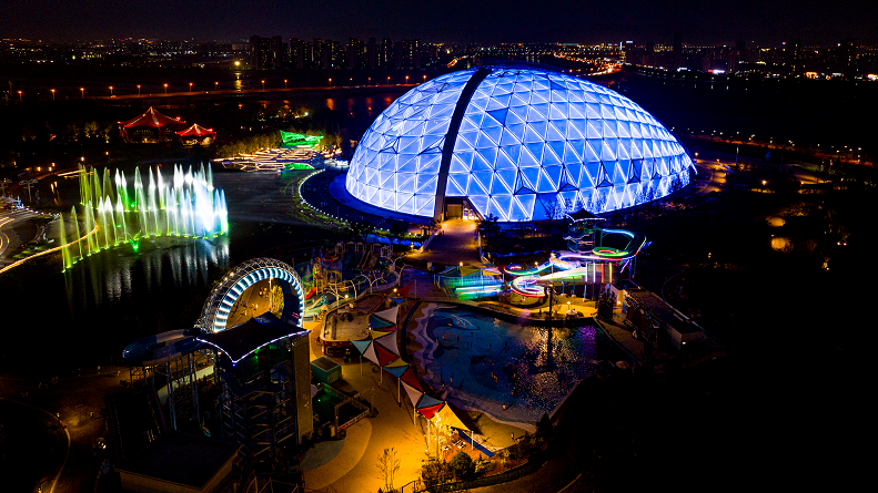 Oriental Eden at night. The egg-shaped biome is illuminated with blue lights which shows its diamond-shaped panels. There are lit fountains in a lake next to the biome and a series of light installations nearby. Lights are reflected in the lake.