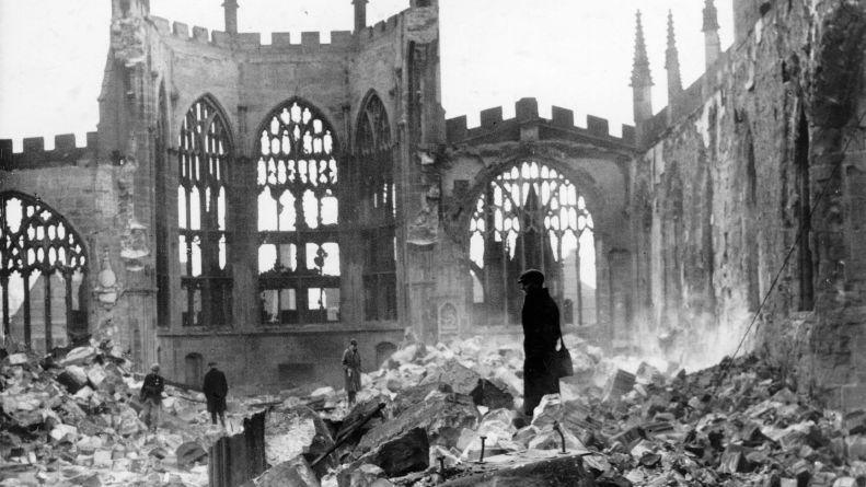 A black-and-white image of people walking through rubble piled up inside a cathedral. The cathedral's end wall remains intact but windows have been blown out.