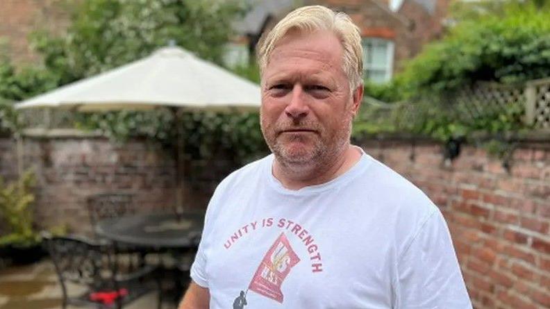 Peter Scarfe, who has blonde hair and is wearing a white t-shirt with the logo "Unity is Strength" in red lettering.