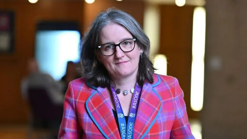 Maree Todd looking right at the camera. She has dark hair. She is wearing dark-rimmed glasses and a blue and purple lanyard around her neck. She is wearing a pink tartan jacket with blue and yellow lines.