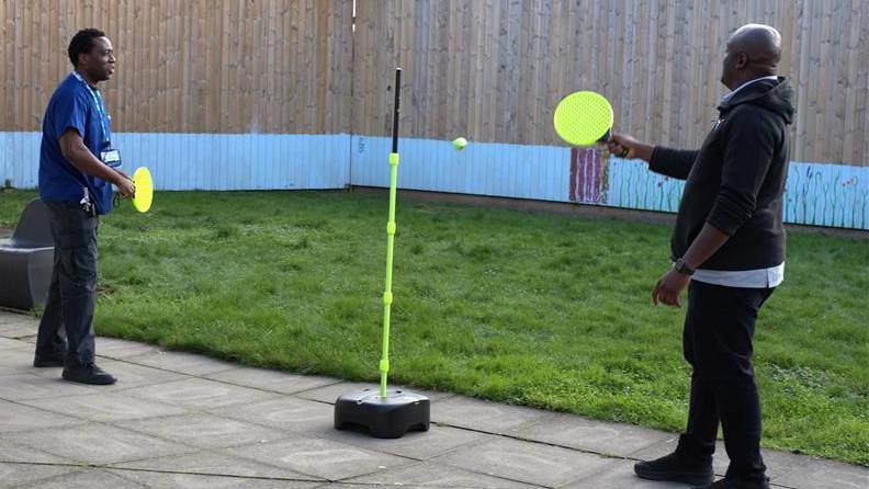 Two men in NHS uniform playing swingball on a paved surface.