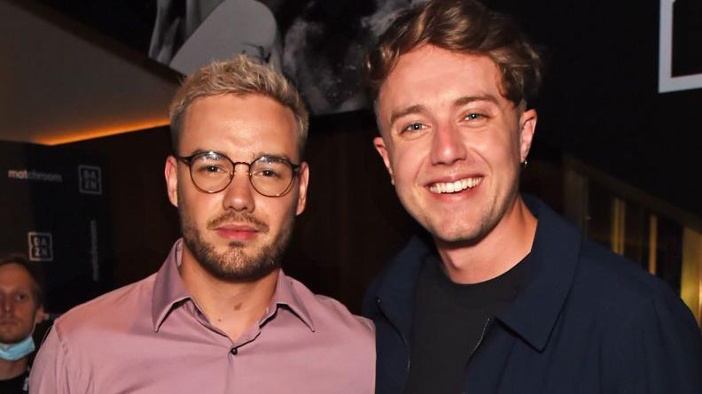 Liam Payne standing with Roman Kemp. Liam is wearing glasses and a pink shirt. He is smiling with Roman who wears a blue jacket and has brown hair