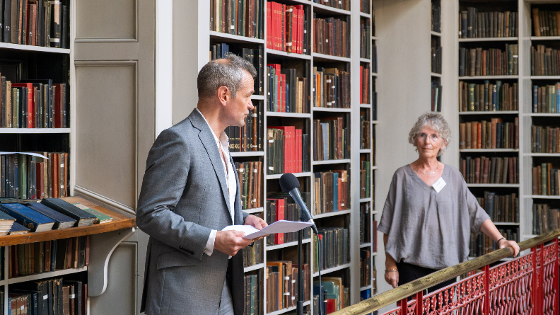 Chairman, Mary Durkin, stands on a balcony with Alexander Armstrong, at the library. Books can be seen behind them.