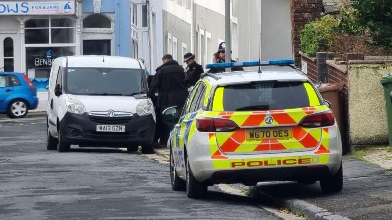 The rear of a police car parked partially on a pavement with a number of police officers in the background and a white van.