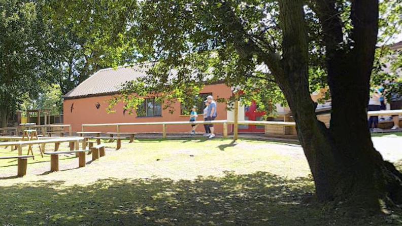 A single-storey brick building set among trees. It has silhouettes of birds of prey on the outside. Two adults and a child are walking by. Also in the picture are some wooden benches.