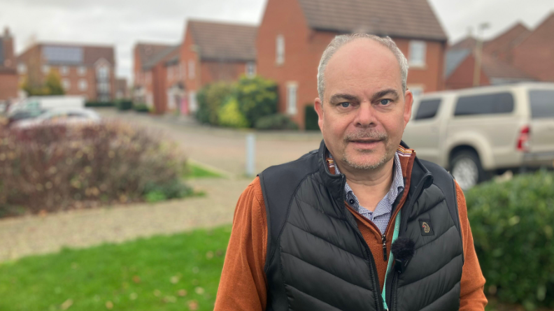 Councillor Ian Snowdon stands in front of red brick houses in Didcot. He is wearing an orange jumper and black gilet.