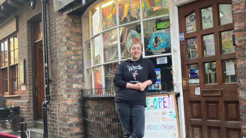 Robyn McGough, from The Portal Bookshop, leans on a fence outside the shop. They are wearing a black hoodie branded with the logo for the rock band My Chemical Romance. 