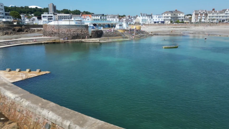 The Havre des Pas lido from a side angle. There is blue and green water with dark patches underneath. There is a grey and red brick wall at the surrounding the seawater pool and in the distance is steps. Behind the lido is other buildings.