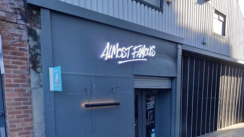 A neon sign for 'Almost Famous' on the front of a grey building above the entrance to the restaurant on Parr Street in Liverpool. 