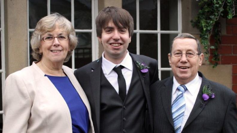 A man wearing his wedding suit, standing with his mother and father either side, in front of the windows of a building.  