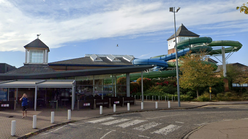 The entrance to Leisure World. It is a modern looking building with a mostly glass facade. However, off to the right is a tower from which a green and a blue flume come out of and lead down towards the back of the building.