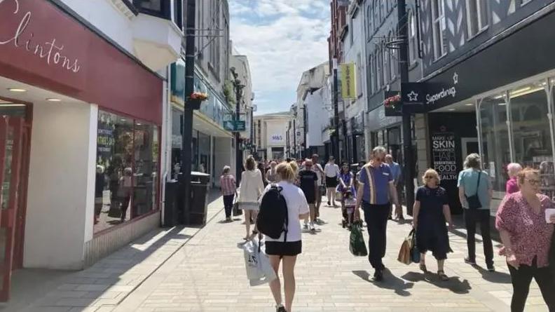 People with shopping bags walking through the high street in Douglas with shops on either side of a paved area. The shops have multicoloured facades.