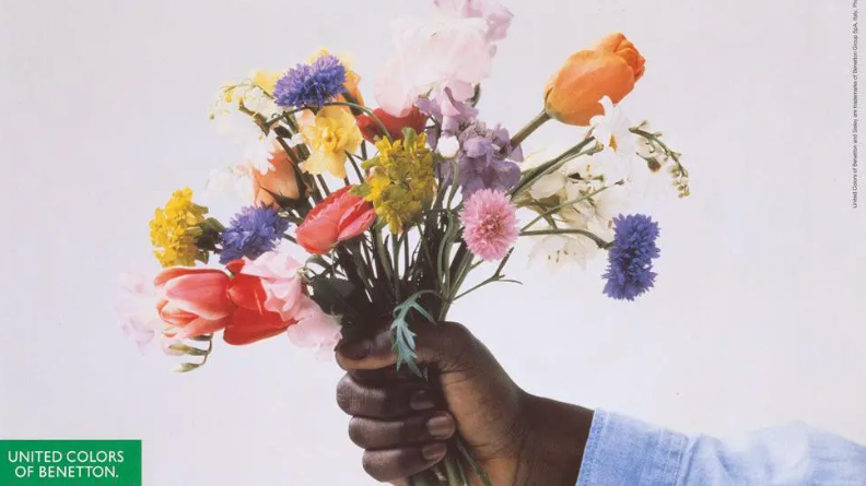 A hand clutches a bunch of flowers including pink and orange tulips, spiky blue cornflowers, pale pink sweetpeas and and white freesias. The green Benetton logo is visible in the bottom left corner. 
