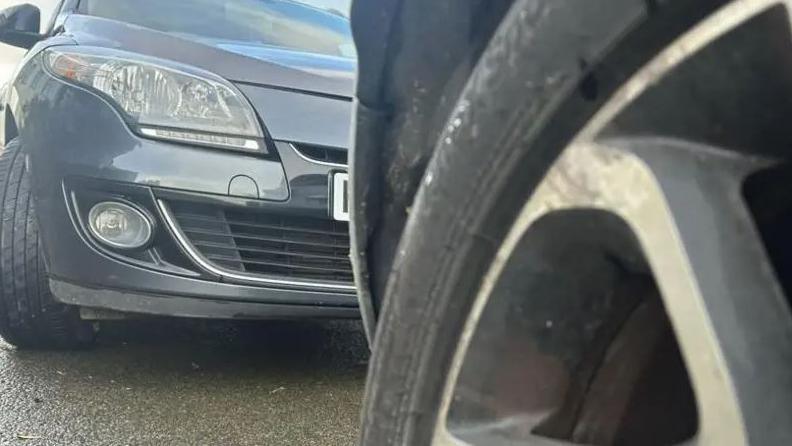 A close-up of a car tyre, with the bumper of a grey car parked on wet concrete behind it.