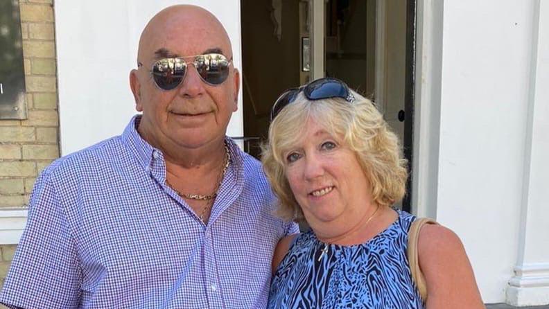 A man and a woman smiling at the camera. He is wearing a blue checked shirt and sunglasses, and she is wearing a blue and black patterned dress with sunglasses on her head