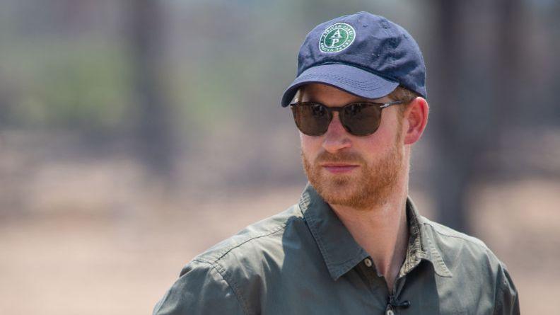 Prince Harry, wearing a blue cap with a green logo in the middle of it and a dark green coloured shirt, looks to the right, while wearing black sunglasses
