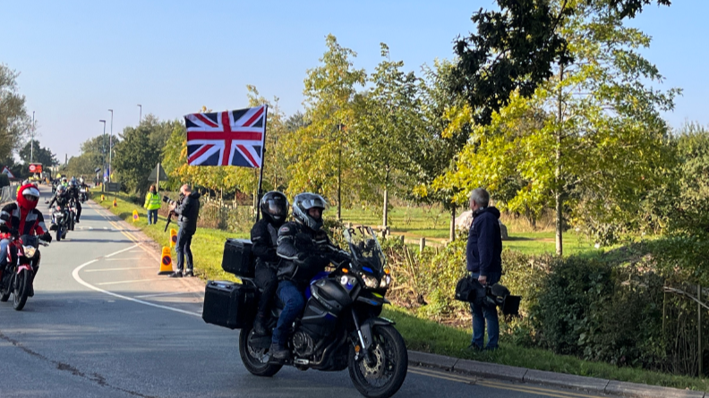 A line of motorbikes, the closest to the camera carries two people, with a union jack flag flying in the wind