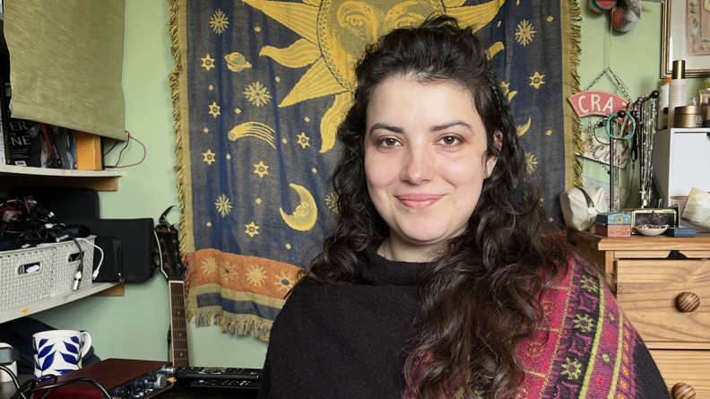 Lily poses for a photograph next to her recording and mixing equipment in her bedroom in Leominster. Behind her are shelves of electronic equipment, a tapestry with the Sun, moons and stars, and a guitar.