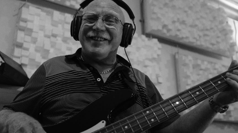 A black and white photo of Alan playing a bass electric guitar with headphones on and smiling to camera