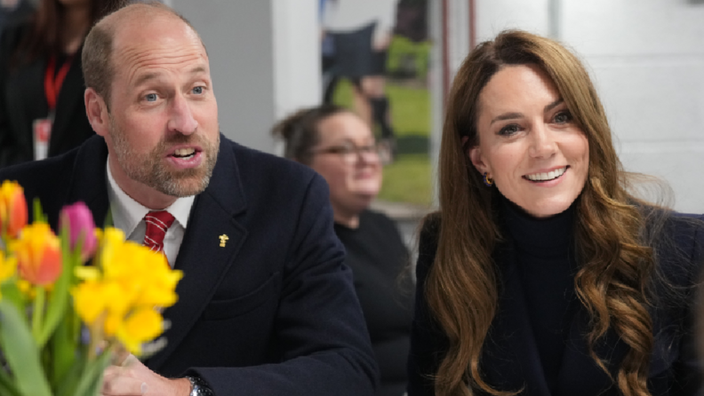 The Prince and Princess of Wales in the Sir Tasker Watkins Suite in the Principality Stadium. Both aren't looking at the camera. William sits on the left, talking to someone off camera, whilst Kate sits on his right, smiling. In the left corner there are a bouquet of flowers, including daffodils. A woman  wearing glasses can be seen in the background.