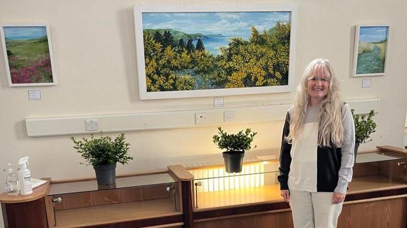 Katrina Bass standing in front of three of her landscape painting, which are hanging on a wall. They have bright colours of yellows, blues and greens. She has long grey hair and is wearing a black, white and grey jumper. There are also pot plants on a shelf underneath the paintings.