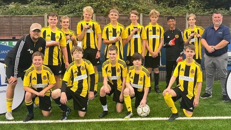 A team of 11 players in a football team, wearing yellow and black kits with their coaches on either side. They are on an artificial football pitch.