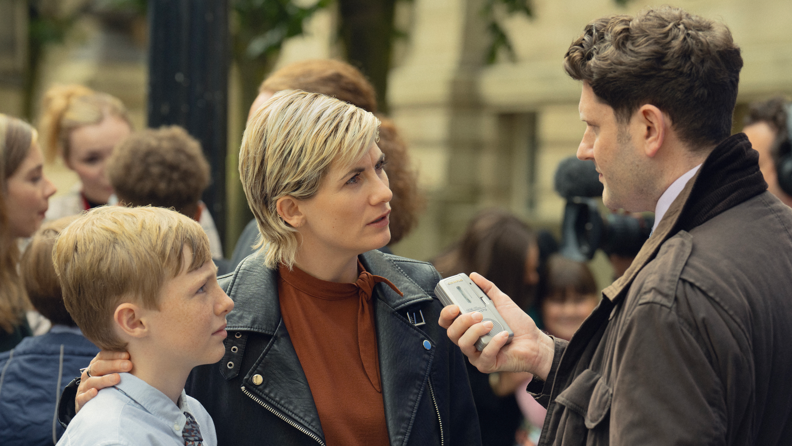 A still picture from a scene in the Toxic Town drama. Jodie Whittaker, in a black leather jacket and brown top, has her arm around a young boy. They are being interviewed by a male reporter in a brown coat who is holding a tape recorder near her mouth. They interview is taking place outside a court building.