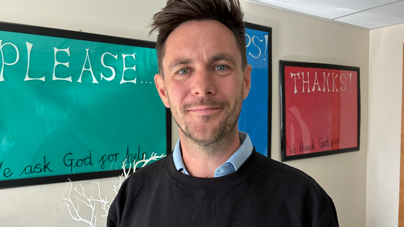 Dave Hall is looking at the camera wearing a jumper with navy blue collared shirt underneath. Behind him are large posters which say "please" and "thanks"on a wall.
