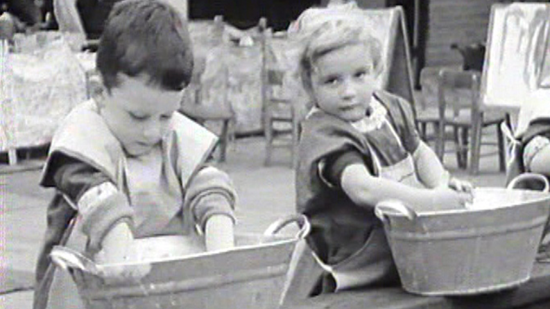 A black and white photo of two young children in an outside nursery setting, playing at washing up in two large bowls