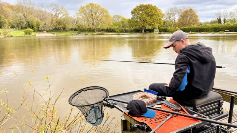  Charlie surrounded by his fishing equipment, sitting on the end of a pier with his fishing rod over the water. He wears a dark hoodie and a grey cap. 