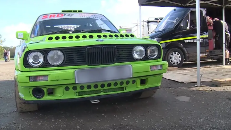 A bright green rally car next to a black van with a blue sky in the background