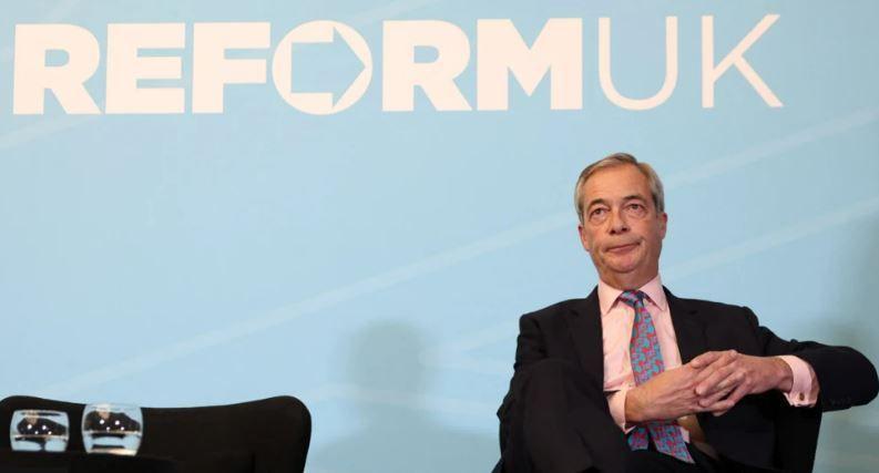 Nigel Farage is seated against the blue background bearing the slogan Reform UK, where a dark suit and pink tie, with his hands clasped together