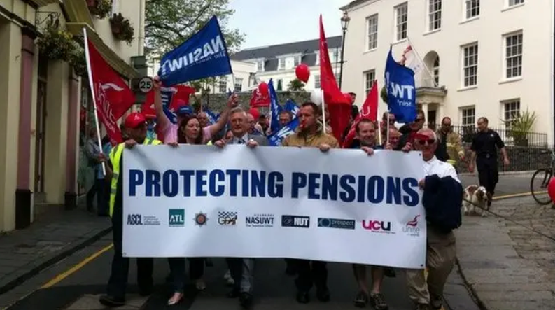 A white banner with the words protecting pensions being held by a number of people at the front of a column of protestors holding banners. 