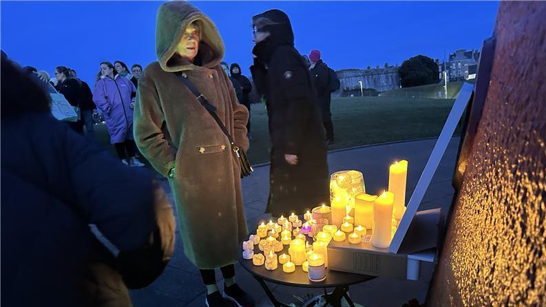 A woman in a thick coat and hood looks at a number of lit candles with other people behind her.