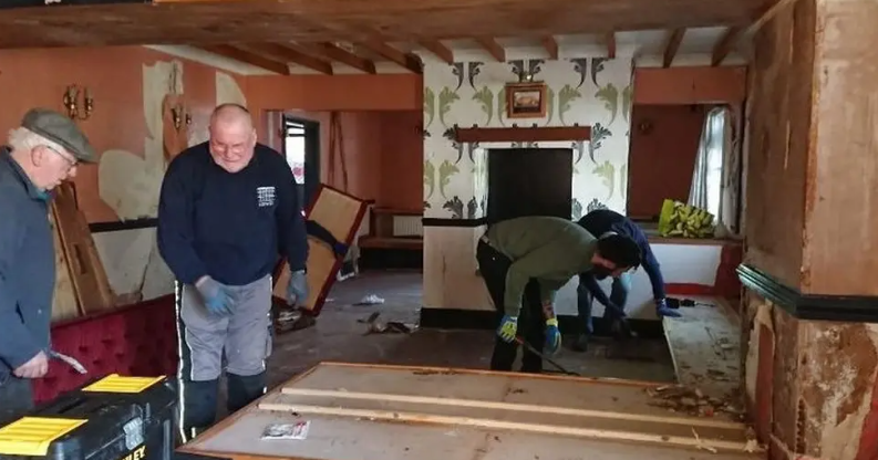 Volunteers wearing work gear pull down parts of the plastering on a wall in the Black Lion pub. The interior is wall-papered pink, with wooden beams and a fireplace.