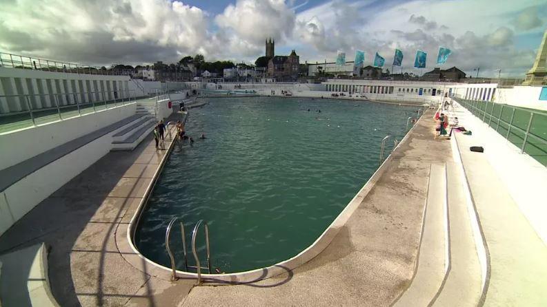 People swimming in Penzance's Jubilee Pool, an outdoor art deco lido. There are clouds in the sky, flags fluttering in the breeze and buildings in Penzance in the background