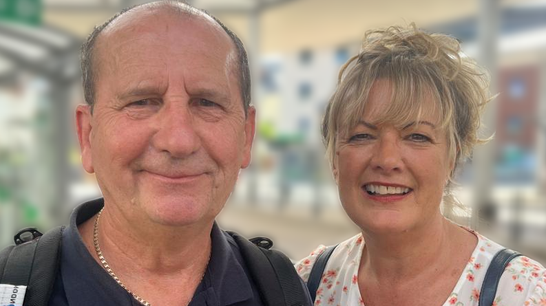 Trevor Turner, left, is looking into the camera smiling. He is wearing a navy top and there are bag straps over his shoulders. Julie Turner, right, is also looking into the camera smiling. She has blonde hair which is tied back. She also has a full fringe. She is wearing a white top with small pink flowers on it. She also has bag straps over her shoulders.