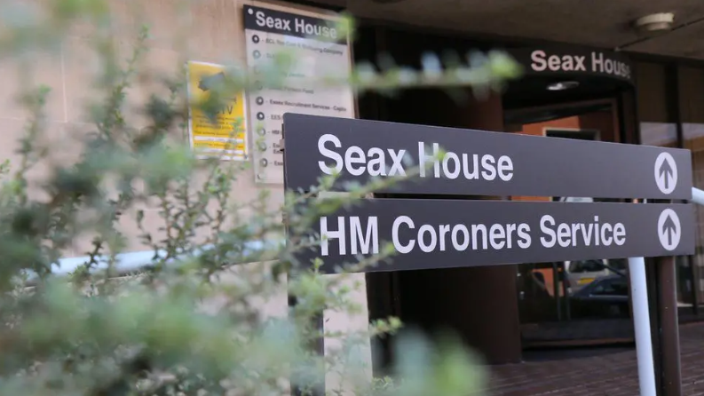 A sign shows the way to Seax House and HM Coroners Service, with an obscured, out-of-focus shrub in the foreground of the picture