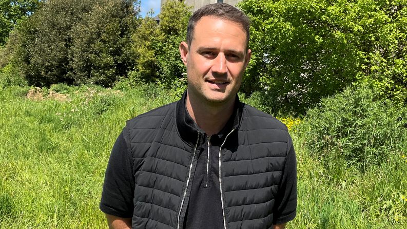 Dressed in a black short sleeved top and gilet Ben Cairney stands in front of a verdant field on a sunny day.