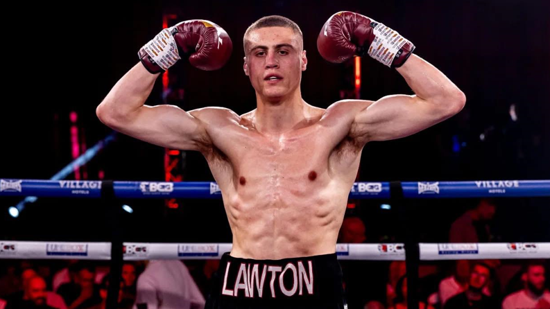 A boxer stands in a boxing ring and raises his gloved hands up to showcase his arm muscles. He has cropped dark hair, and the name Lawton is picked out in letters across the front of his shorts. People can be seen in the darkly lit background sat in seats surrounding the ring. 