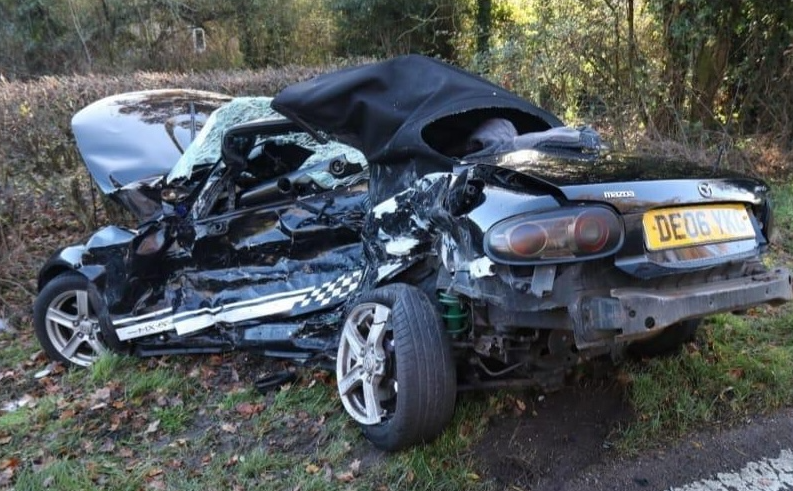 A very badly damaged car, with a smashed windscreen, the passenger side bent inwards and the rear tyre off the wheel. The car is pointing into a hedge