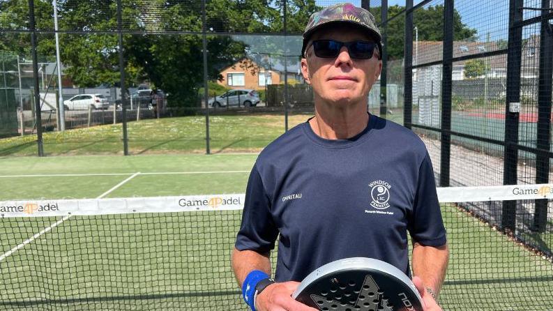 Roger Wheatley, wearing a baseball cap and sunglasses, stands in front of a net. He has a padel racket in his hands and is wearing a blue shirt and wristband.