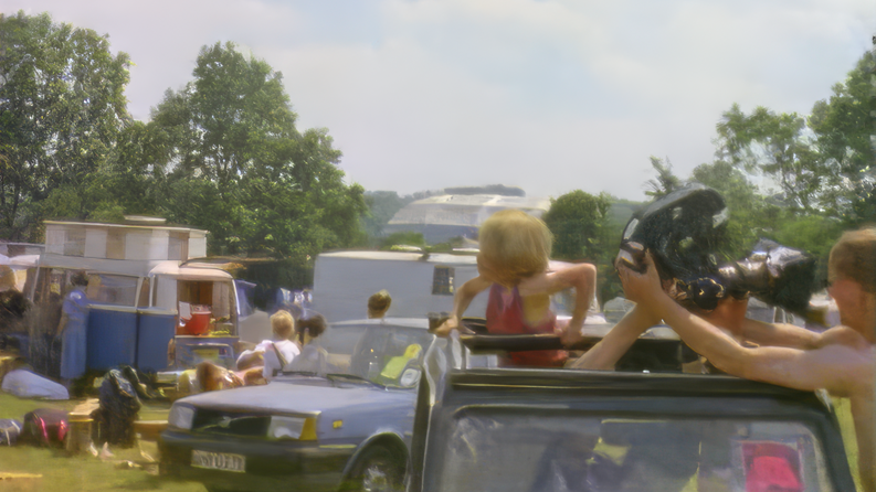 A vintage image of a field with a jeep in the foreground with an open top and someone passing an old-fashioned camera to crew outside. A woman is standing up in the jeep and looking behind her. There is also a blue car from the 90s and some vans in the background with a few festivalgoers milling around. The sky is blue with wispy clouds.