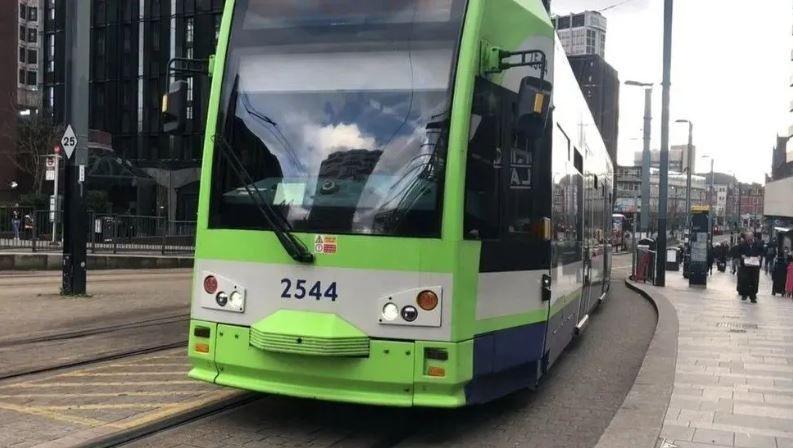 A light green Croydon Tramlink tram along the high street with the grey pavement on the righthand side.