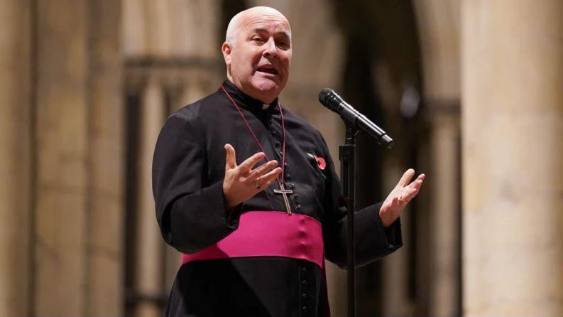 Stephen Cottrell, Archbishop of York, giving a sermon dressed in black with a red belt, wearing a poppy and a cross necklace. He is balding with white hair.