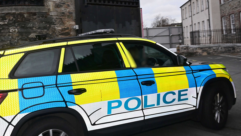 Shows a police car in yellow blue and white colours with the word "police" on the side entering a court complex. 